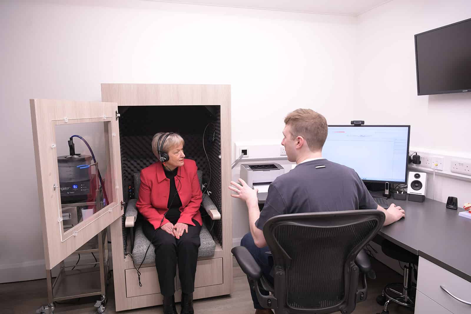 Audiologist discussing a hearing test with a female customer sat in a sound proof booth